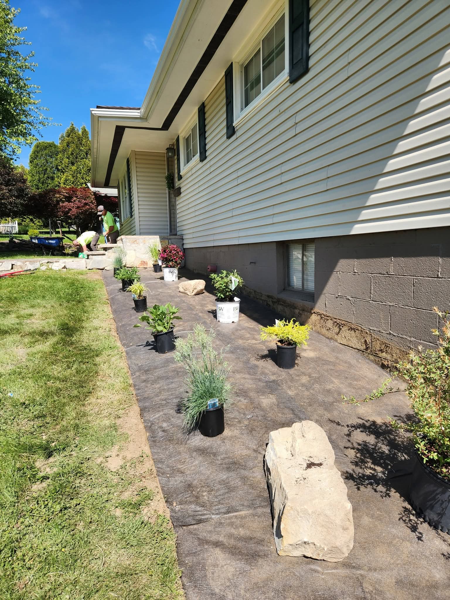 Potted plants laid out on landscape fabric along a house while the crew works