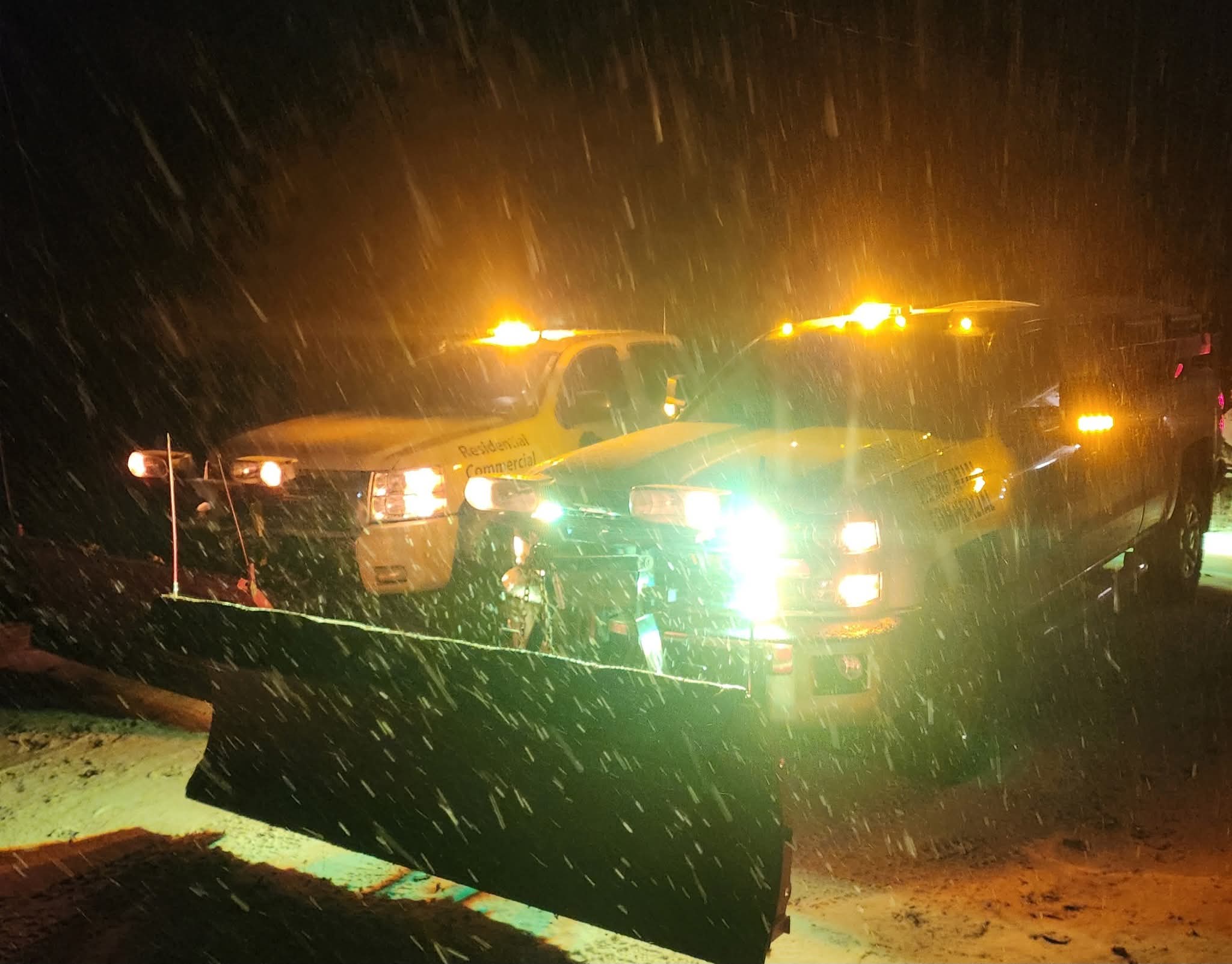 Two plow trucks with amber lights in falling snow at night